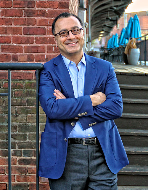 Yasser Youssef standing in front of the historic Railroad Building in downtown Winston-Salem, smiling and wearing a royal blue blazer.